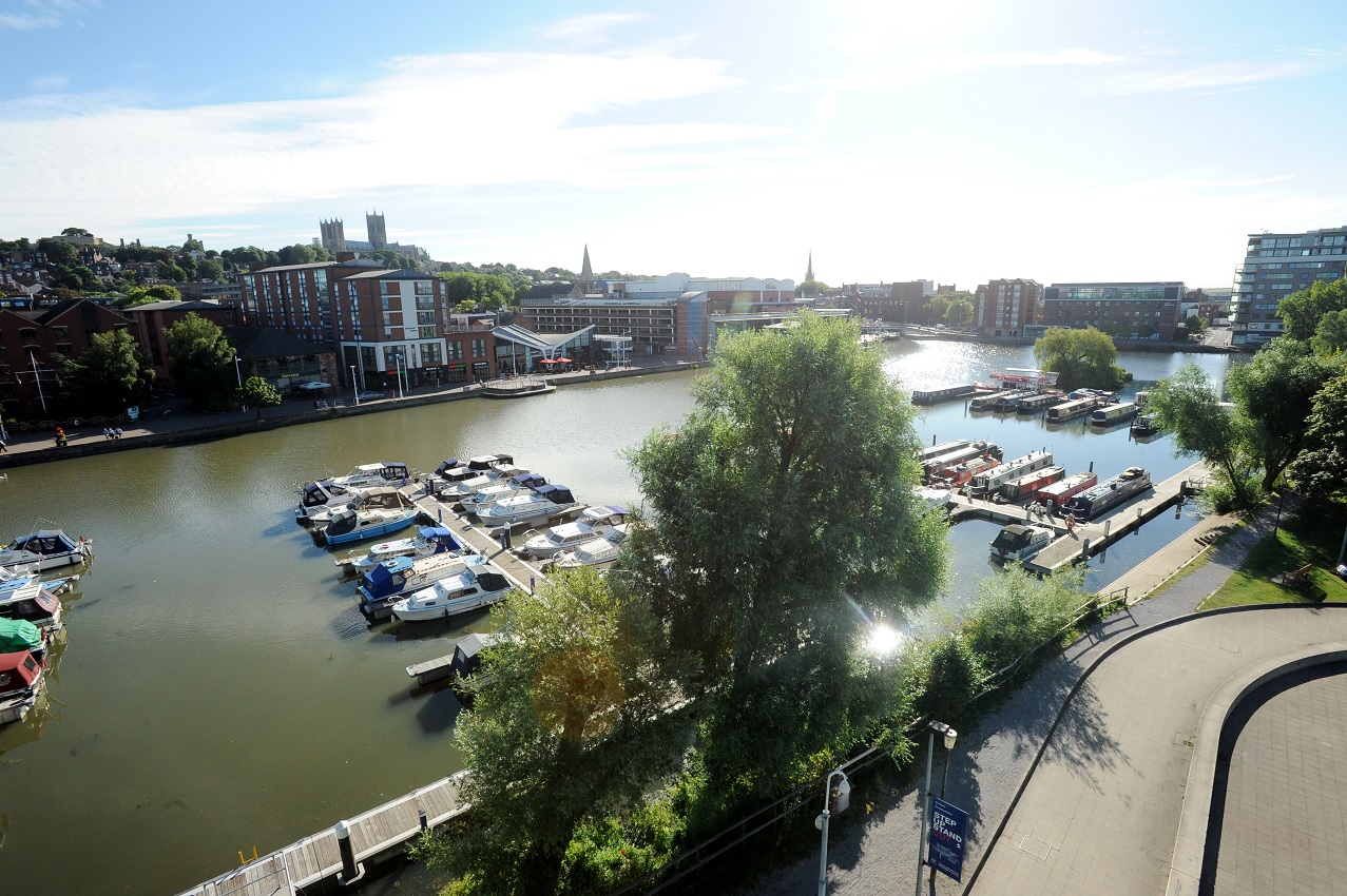 Landscape photograph of the Brayford Pool in Lincoln. Features boats in the water and several buildings and trees around the water.