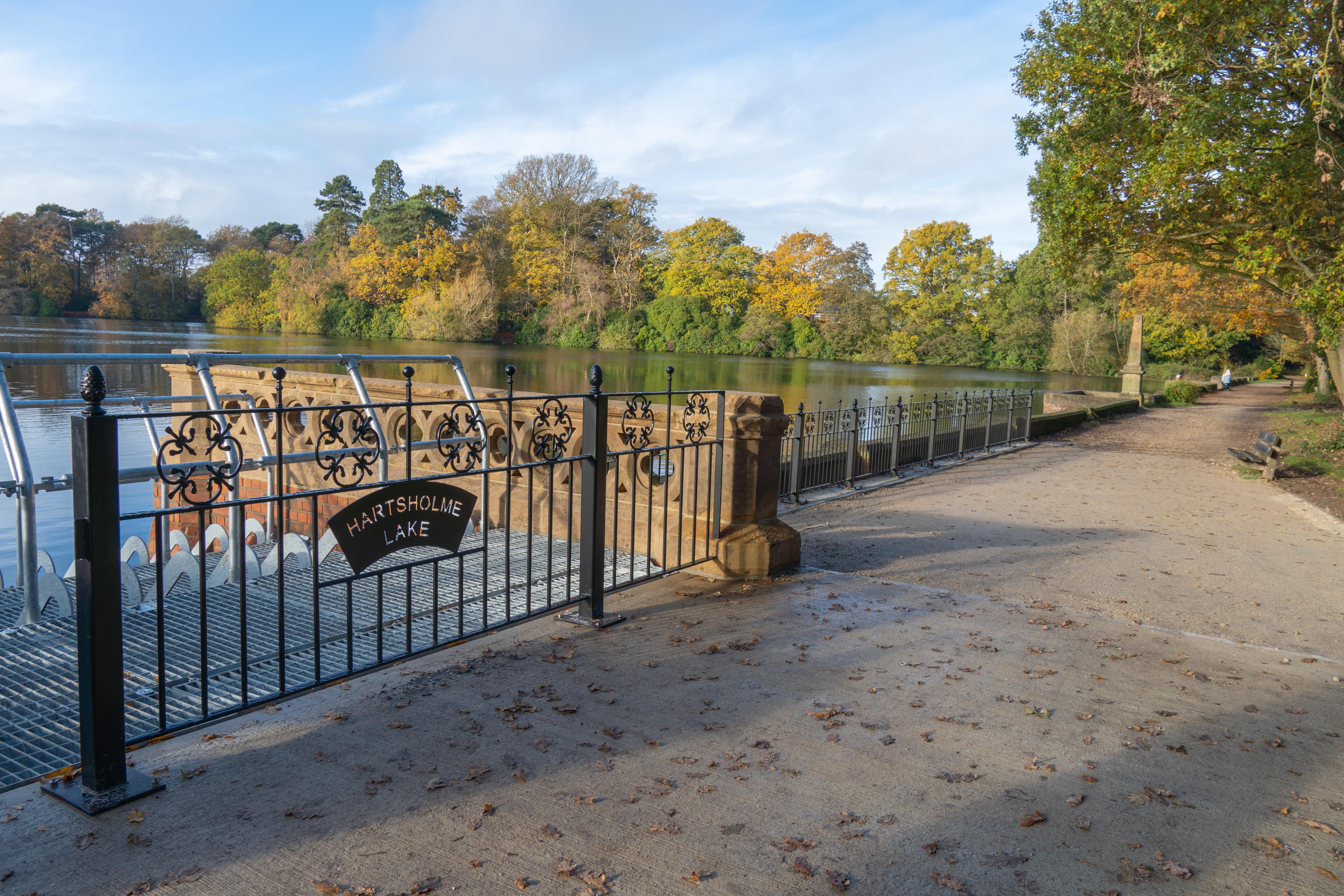 A lake with trees in the background. In the foreground there are some railings with the words ‘Hartsholme Lake’.