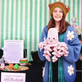 A young teenager stands in front of her stall wearing a crochet hat, holding a handmade crochet octopus plushie. A sign in front of the stall displays the business name, 'Off the Hook'