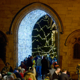 A glowing silver light installation that illuminates Exchequer Gate, close to Lincoln Cathedral. The Tree of Life decorated with gold fairy lights is seen on the other side of the gate.