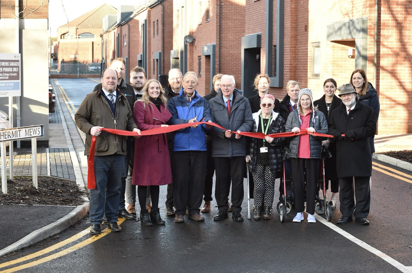 A group of people stood holding a red ribbon on a new street.