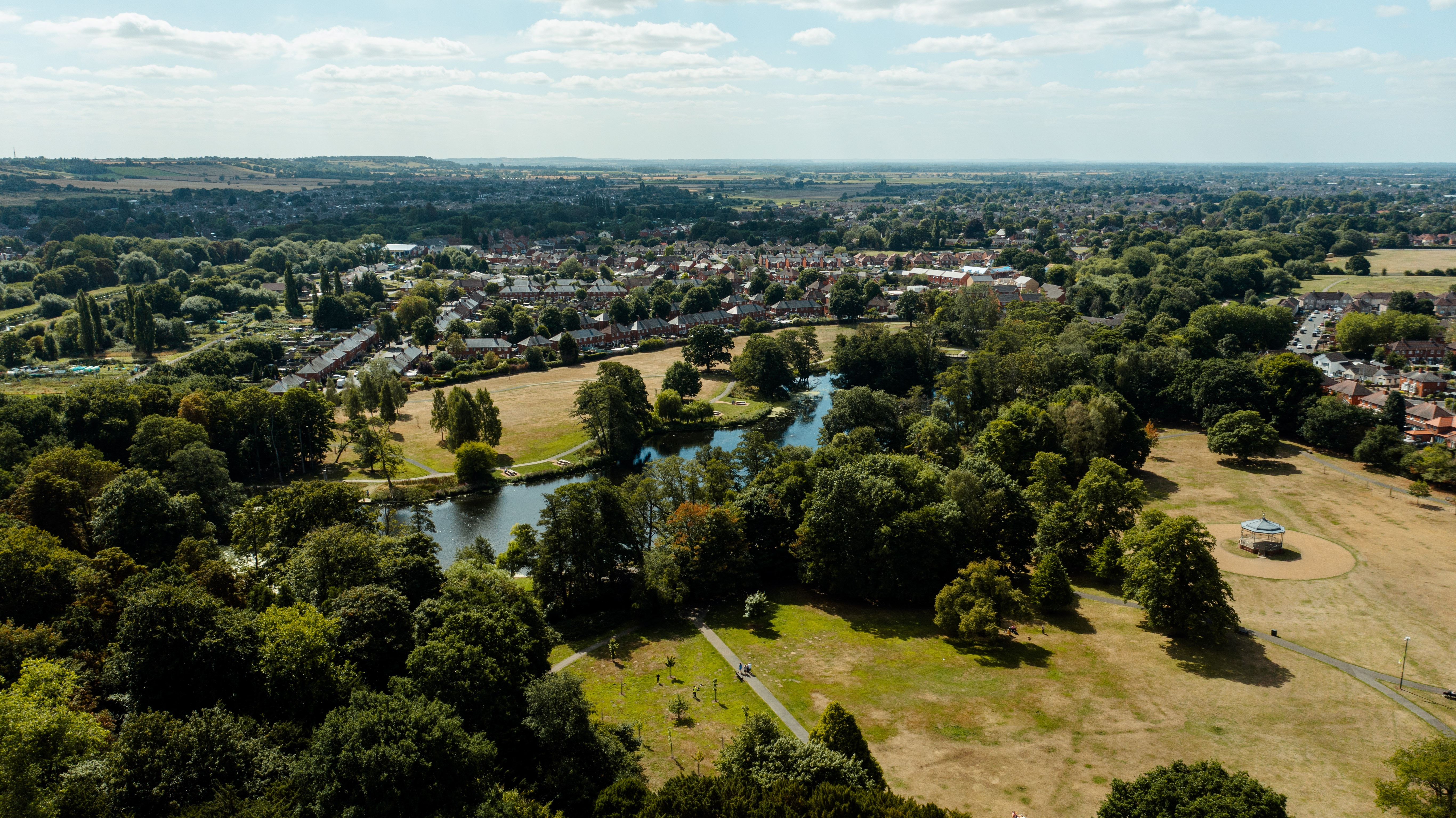 A drone image looking down on the lake at the edge of a green park surrounded by trees.