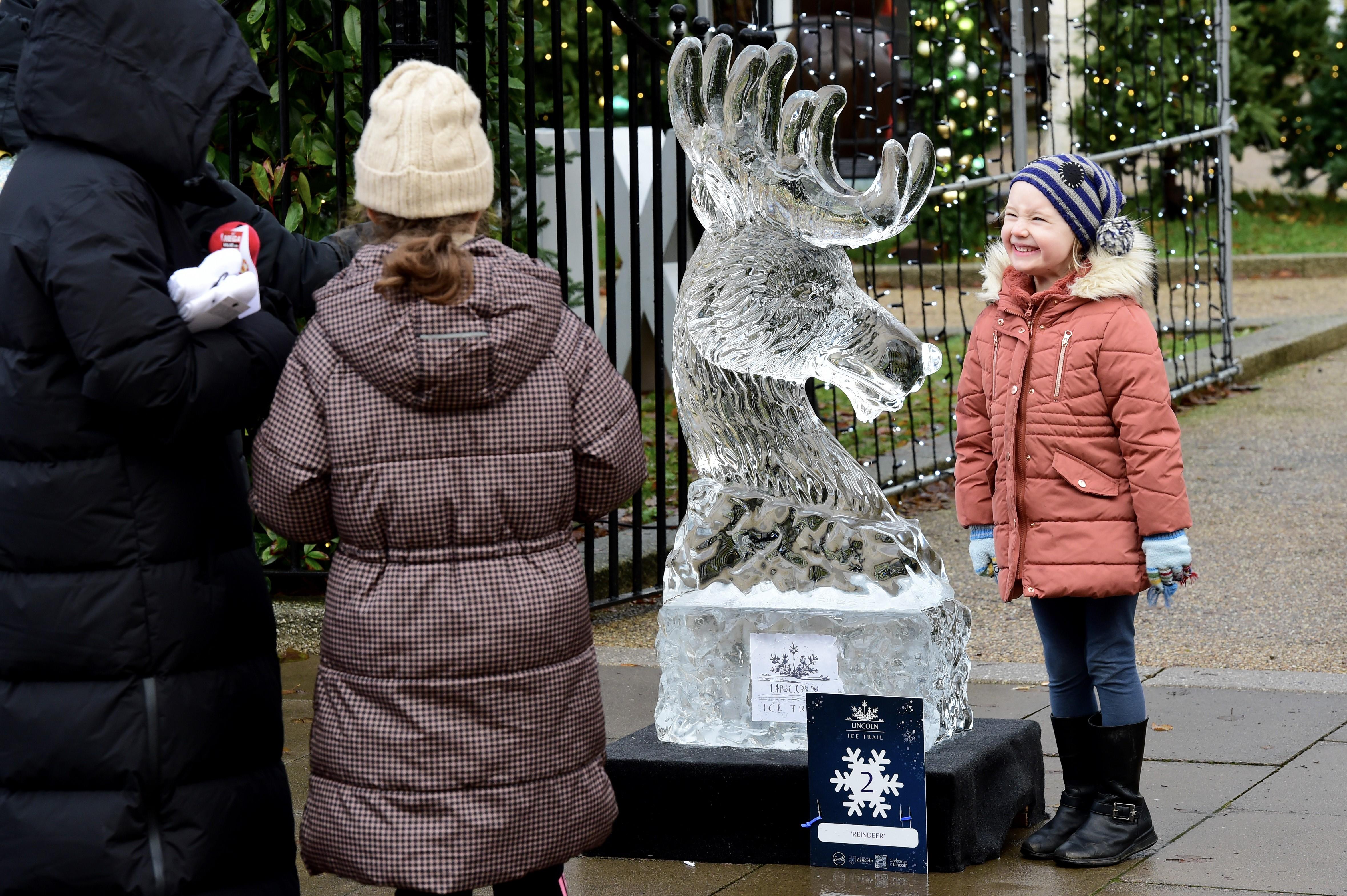 A young girl is smiling as she stands next to an ice sculpture of a reindeer. The girl is wearing a coat, woolly hat and gloves and there are twinkling Christmas lights which can be seen in the background.