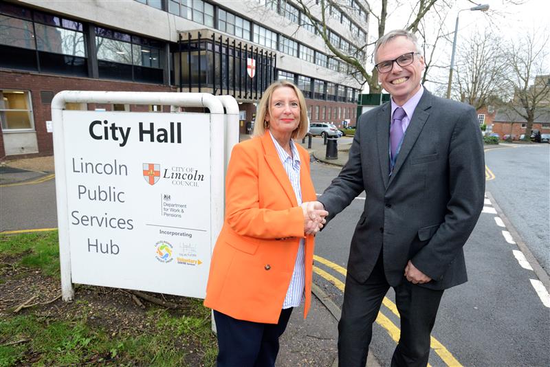 A woman dressed in an orange blazer shaking hands with a man in a suit in front of the City Hall sign.